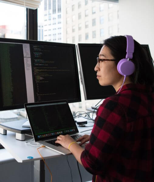 Woman wearing purple headphones while typing on laptop and looking at 2 desktop computers working on devops services.